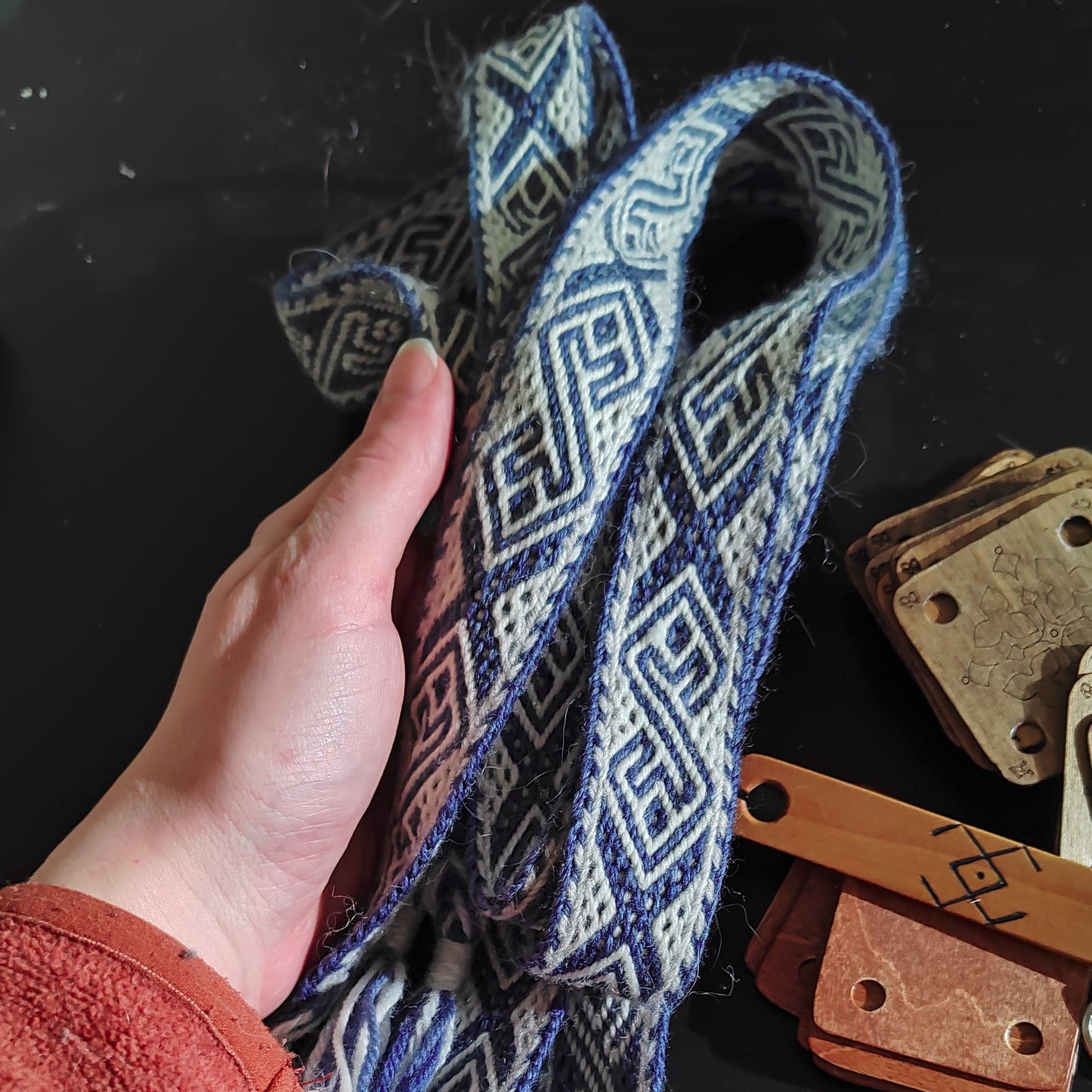 A person's hand holding a woven textile with a geometric pattern, alongside weaving tools on a table.