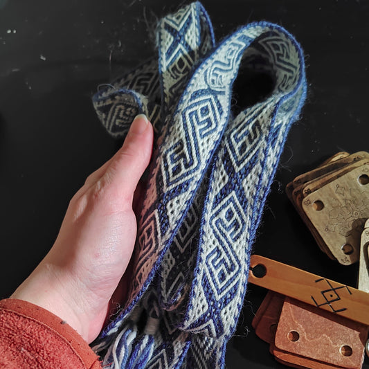 A person's hand holding a woven textile with a geometric pattern, alongside weaving tools on a table.